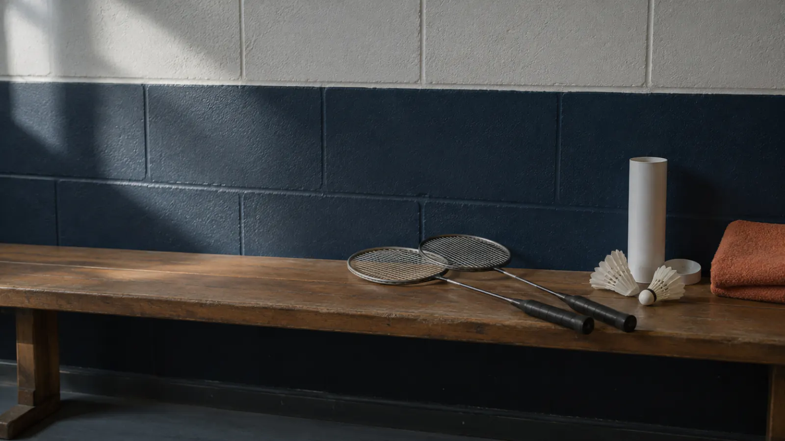 Two badminton rackets, a tube of shuttlecocks and an orange towel on a wooden bench against a navy wall